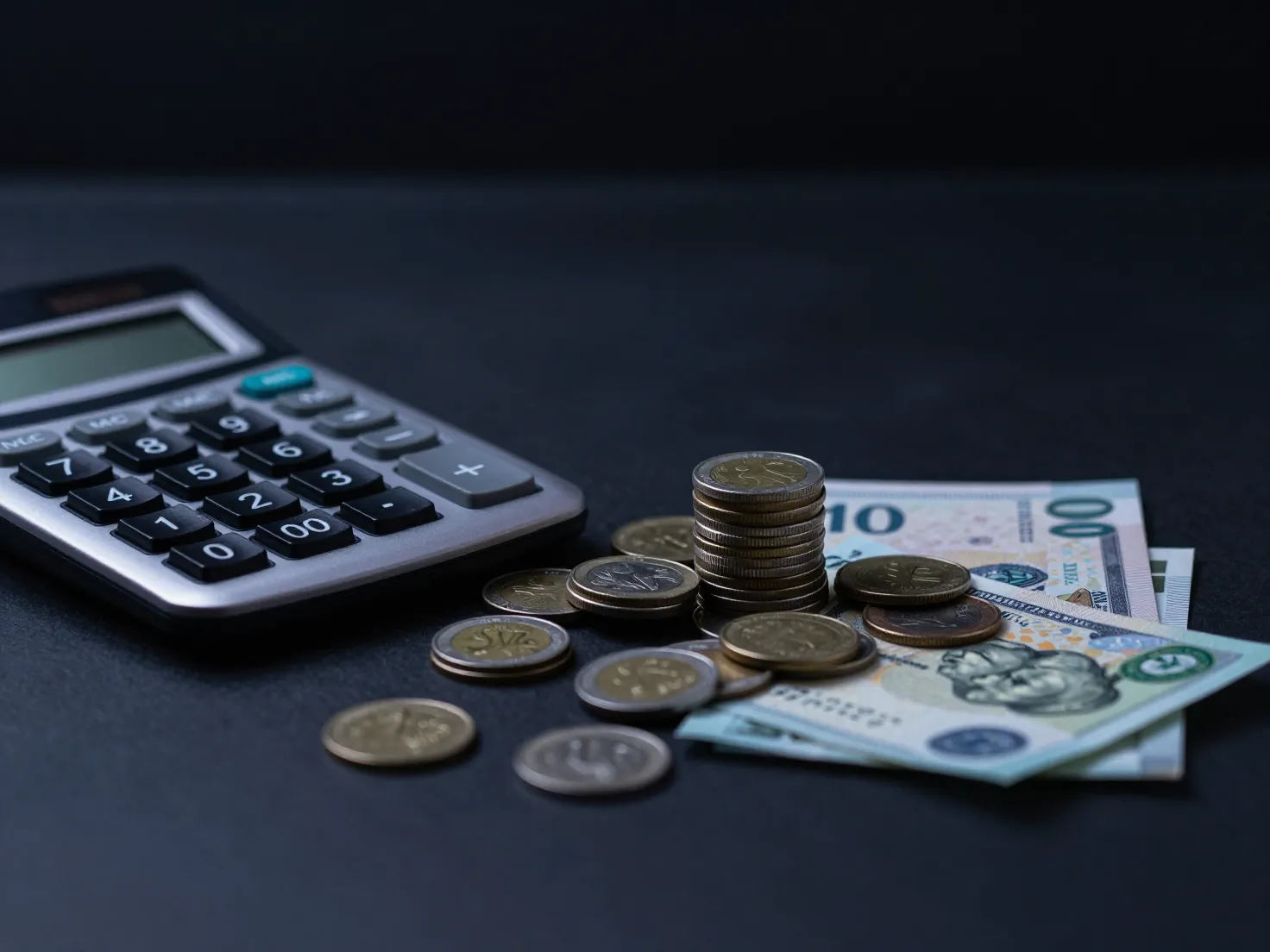 A calculator with coins and banknotes on a dark desk